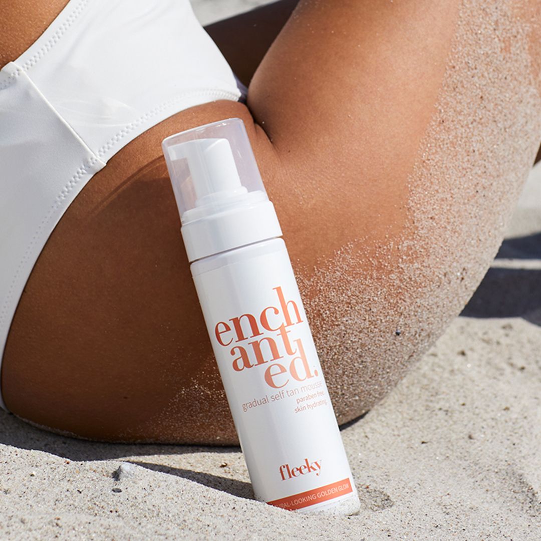 Weißes Mousse-Behältnis auf Sand. Aufschrift: enchanted. Gradual Self Tan Mousse. Marke: fleeky. Gebräunte Haut.