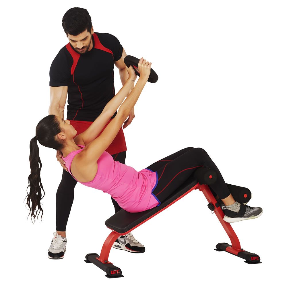 Person trainiert auf einer roten und schwarzen Mini-Ab-Bench. Eine Person hilft. UFC-Logo auf der Bank. Frau in Sportkleidung, Mann in Sportkleidung.