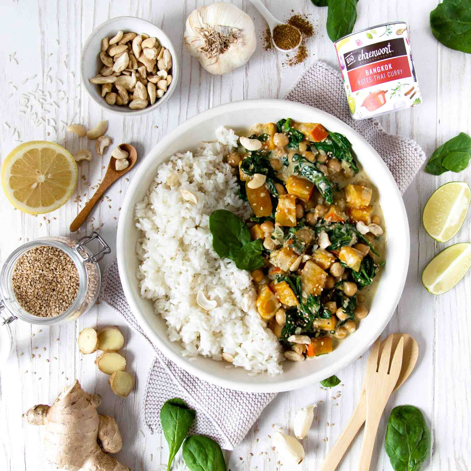 Rotes Thai Curry mit Reis, Spinat, Süßkartoffeln und Cashewkernen. Zutaten wie Ingwer, Knoblauch und Limette sind arrangiert.