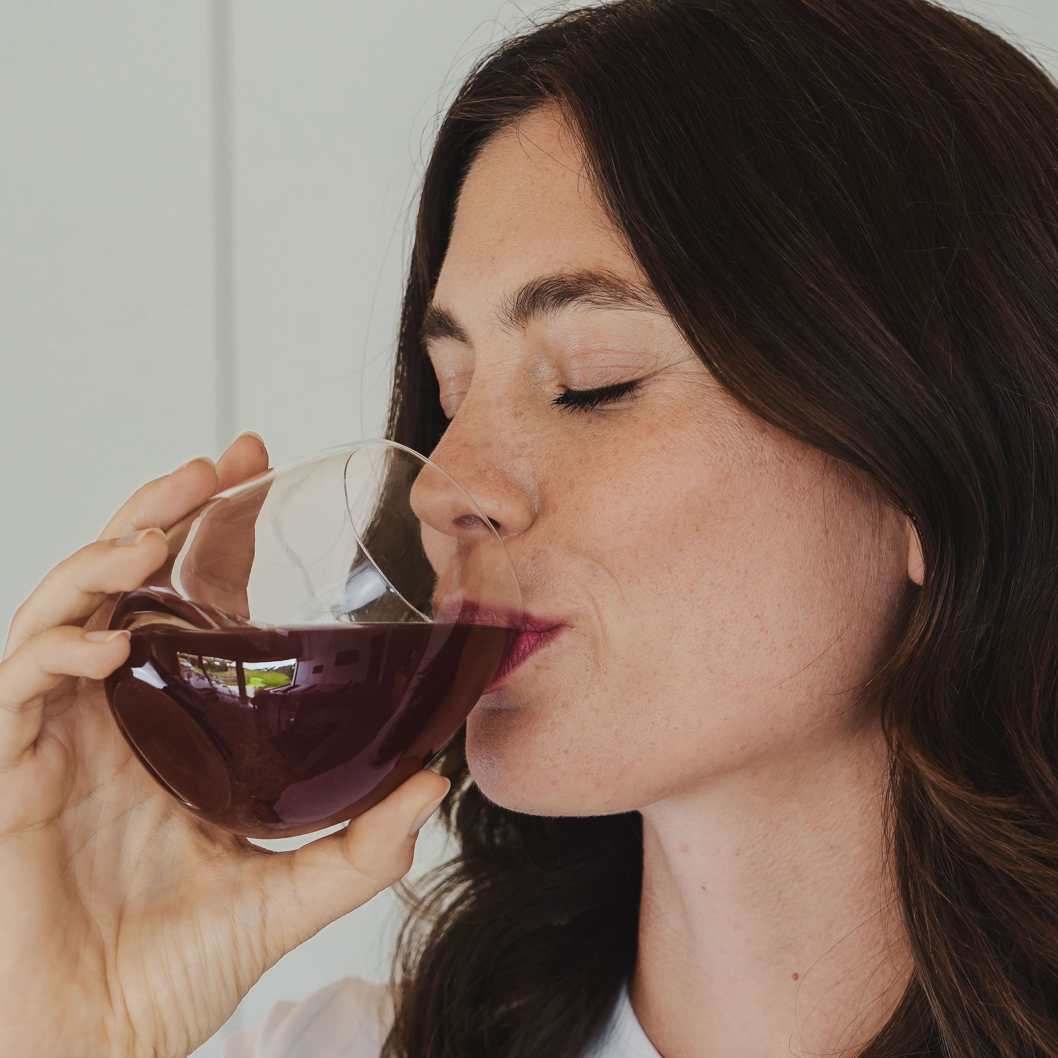 Frau trinkt aus einem Glas. Flüssigkeit ist rot. Sie hat dunkle Haare und schließt die Augen.