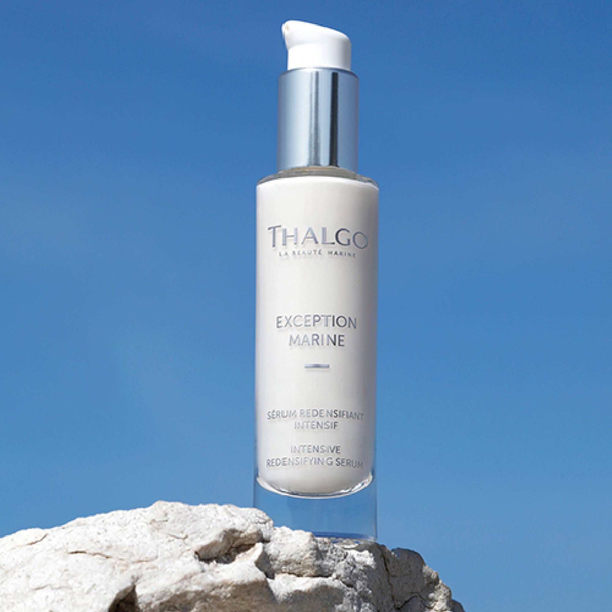 Serumflasche mit silbernem Pumpkopf. Aufschrift: THALGO, EXCEPTION MARINE, Serum Intensif. Steht auf einem Felsen vor blauem Himmel.