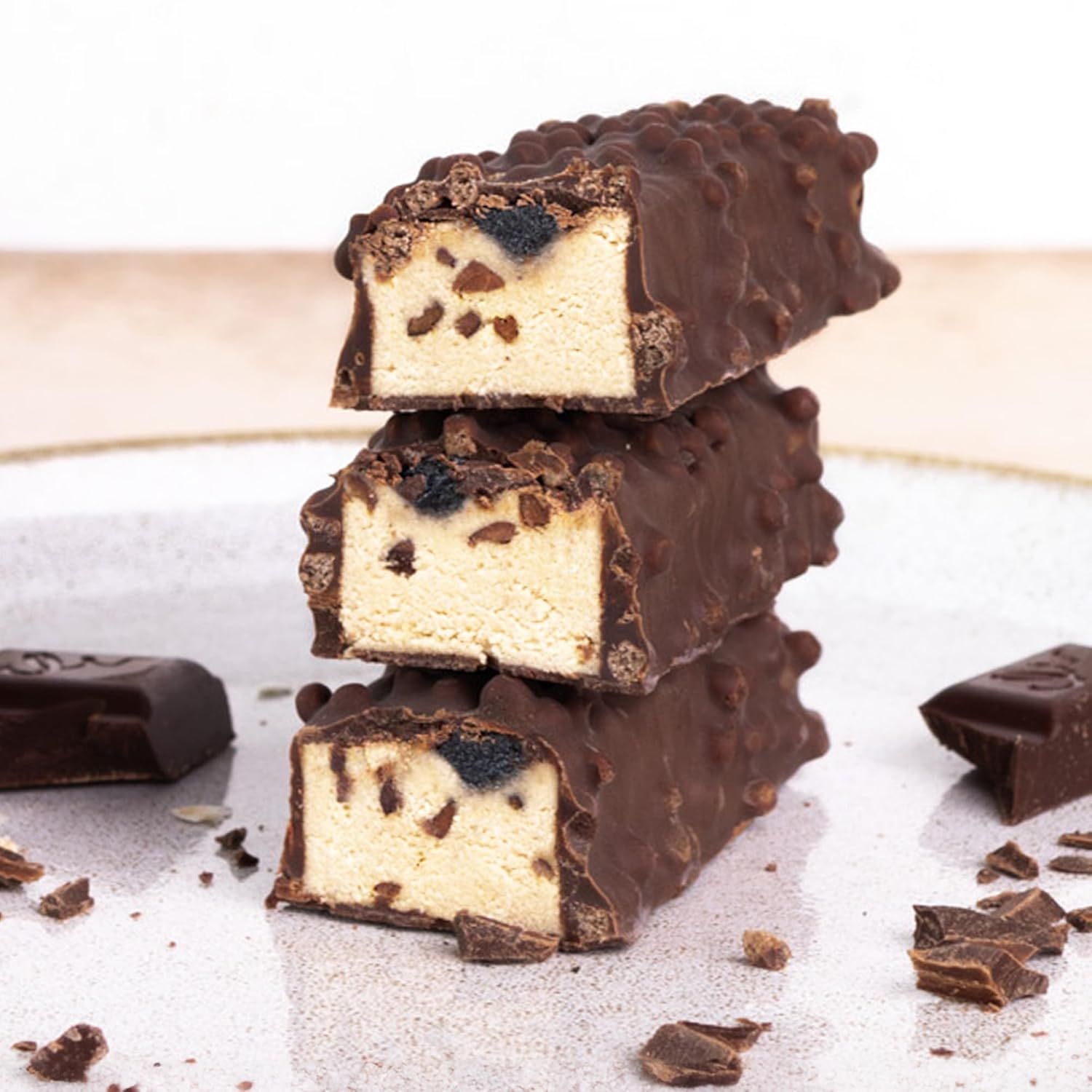 Drei aufgeschnittene Proteinriegel, mit Schokoladenüberzug und Füllung. Auf einem Teller. Einige Schokostücke liegen daneben.