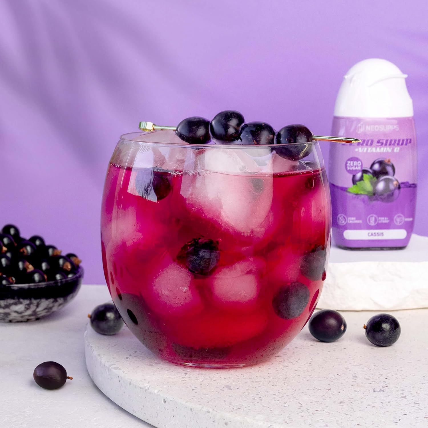 Glas mit Getränk, Eiswürfeln und Johannisbeeren. Daneben eine Flasche Neosupps Zero Sirup Cassis.