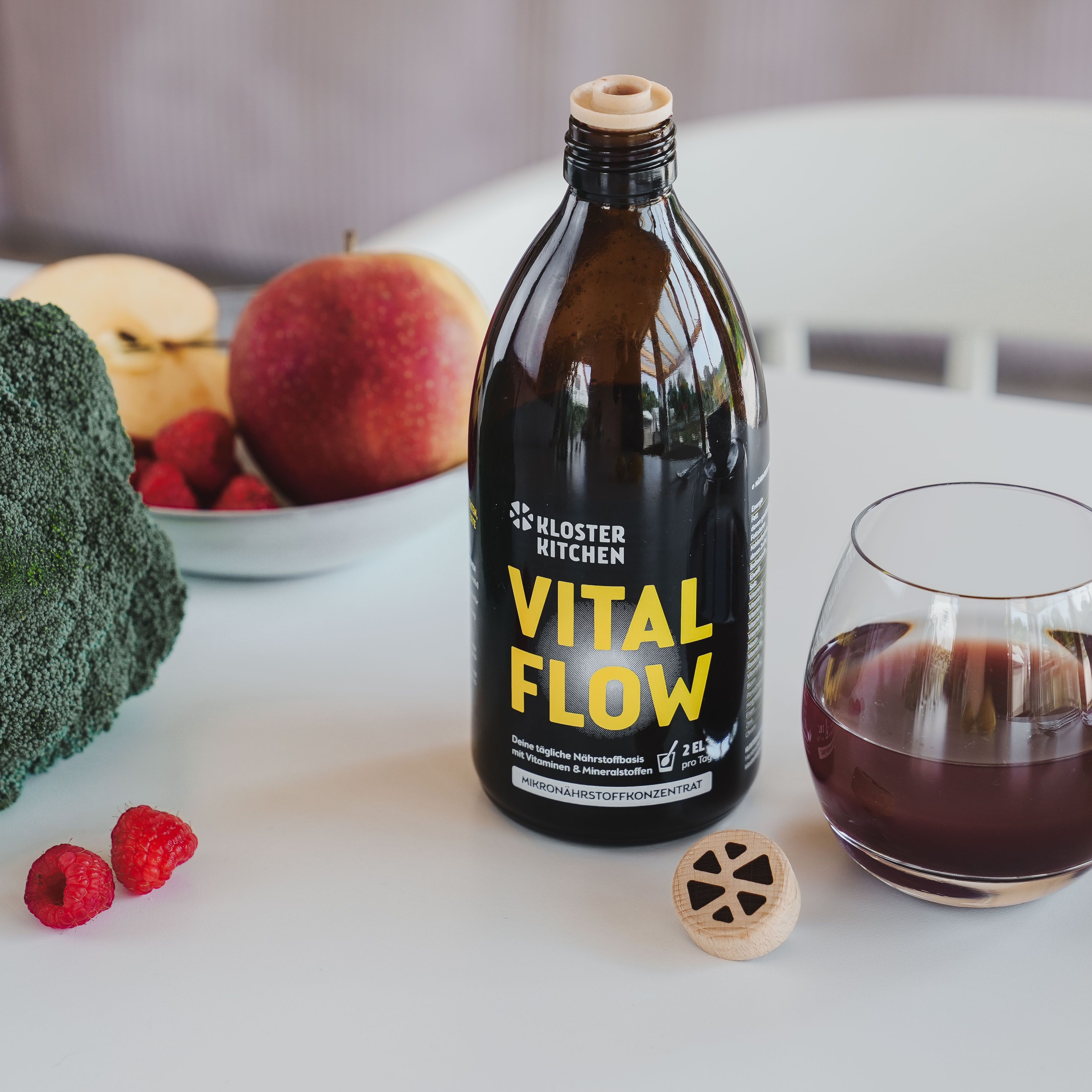 Flasche VITAL FLOW, daneben Glas mit Flüssigkeit, Himbeeren, Apfel, Brokkoli. Auf Tisch.