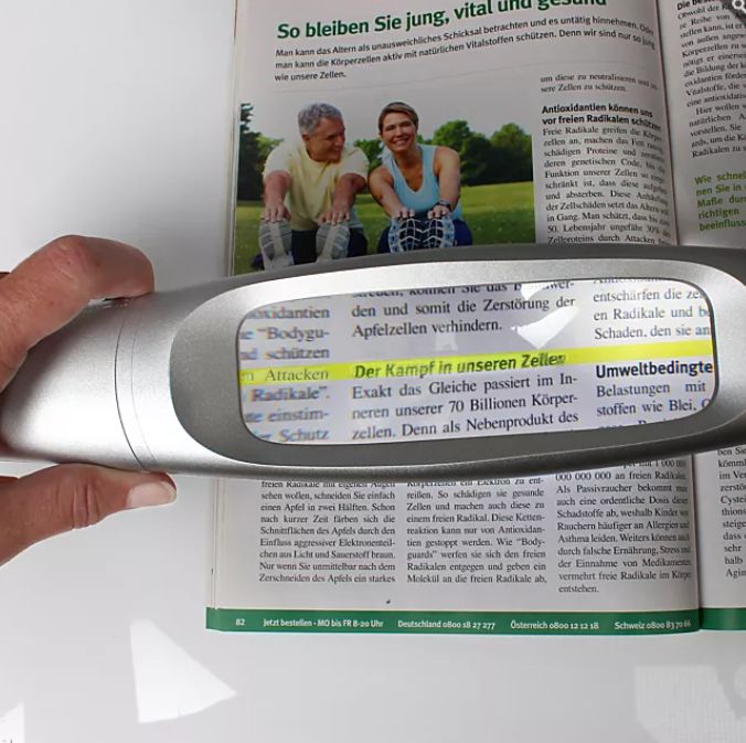 Hand, die LED-Lupe über einer geöffneten Zeitschrift hält. Gehäuse silberfarben, rechteckige Linse. Text wird vergrößert.