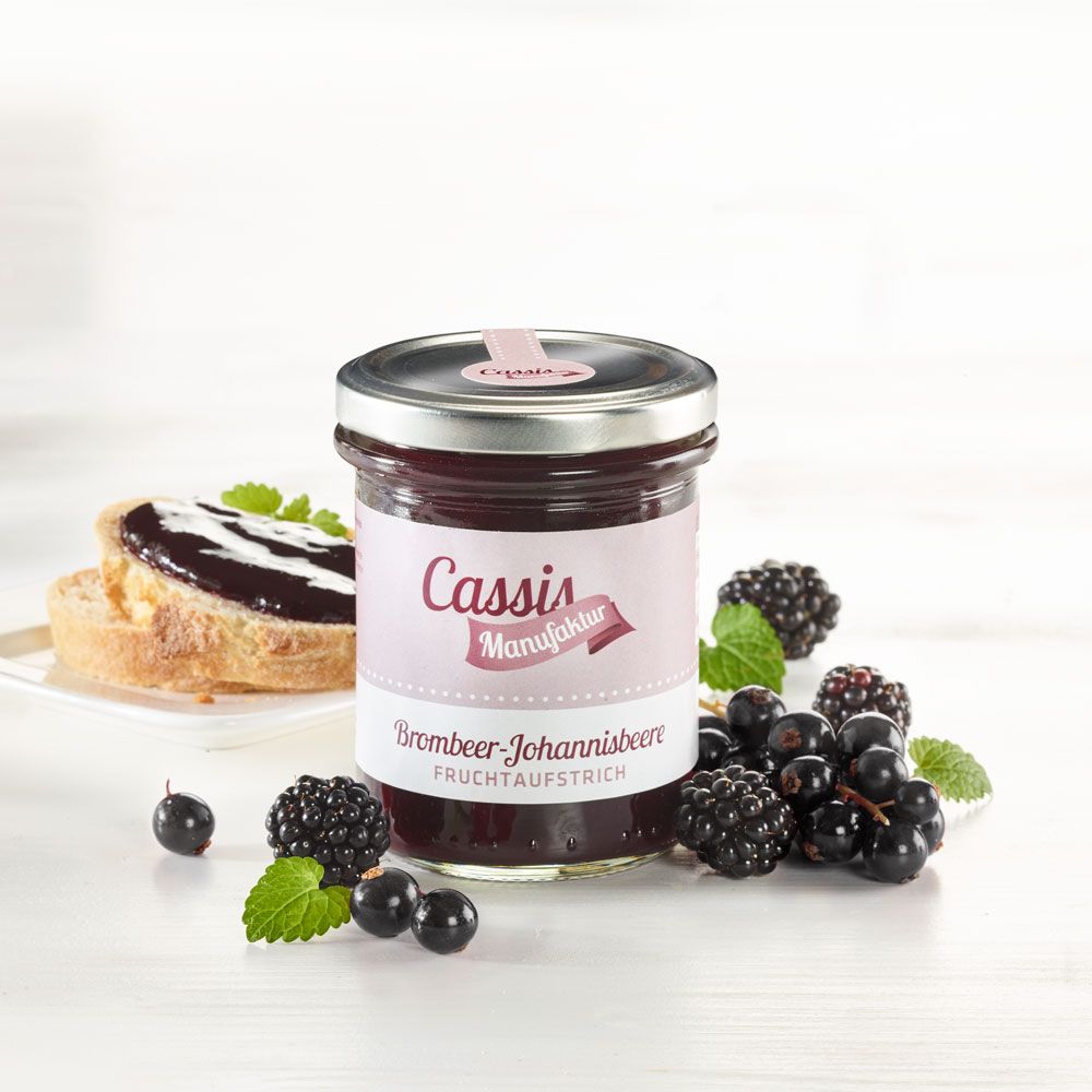 Glas Fruchtaufstrich Brombeer-Johannisbeere, neben Brot, Beeren und Minze. Etikett "Cassis Manufaktur".