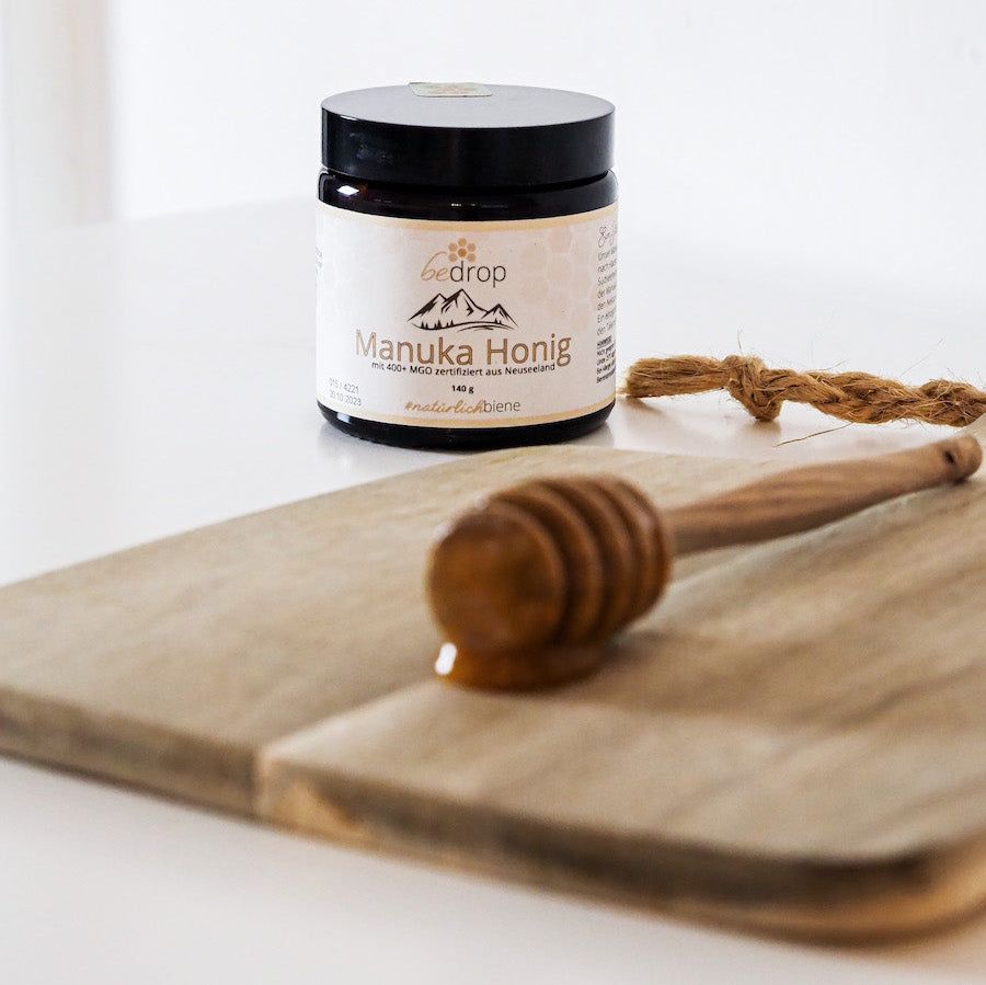 Braunes Glas mit Manuka Honig-Etikett. Aufschrift: #natürlichbiene. Auf einem Holzbrett liegt ein Honiglöffel.