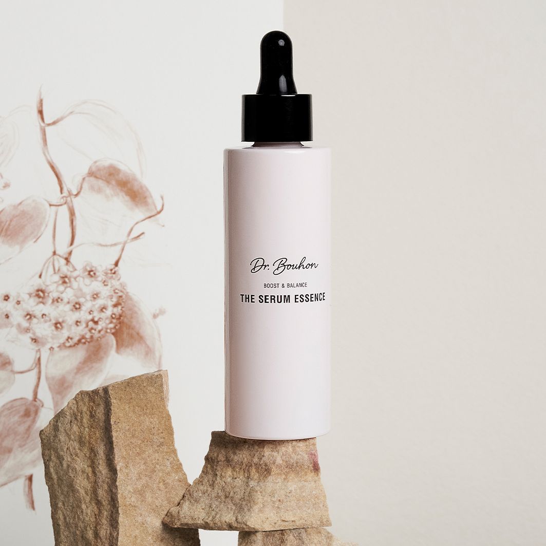 Flacon rose avec bouchon noir sur des pierres. Inscription : Dr. Bouhon, THE SERUM ESSENCE. Fond : floral.