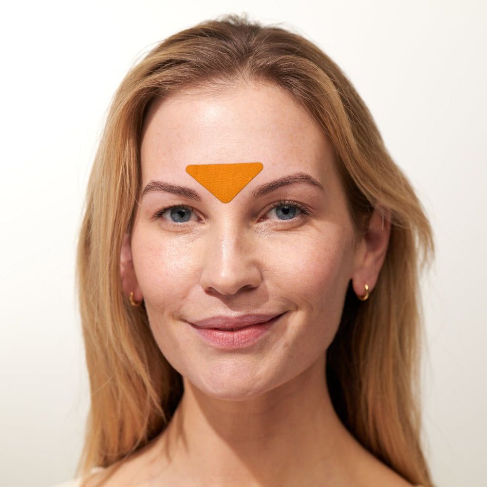 Porträt einer Frau mit einem orangefarbenen Gesichtspatch auf der Stirn. Sie trägt Ohrringe und lächelt.