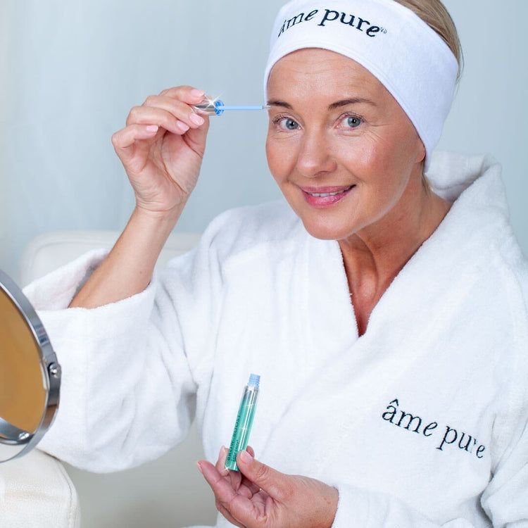 Frau im Bademantel mit Augenbrauenserum. Aufschrift: âme pure. Frau trägt Stirnband. Serum mit Applikator.
