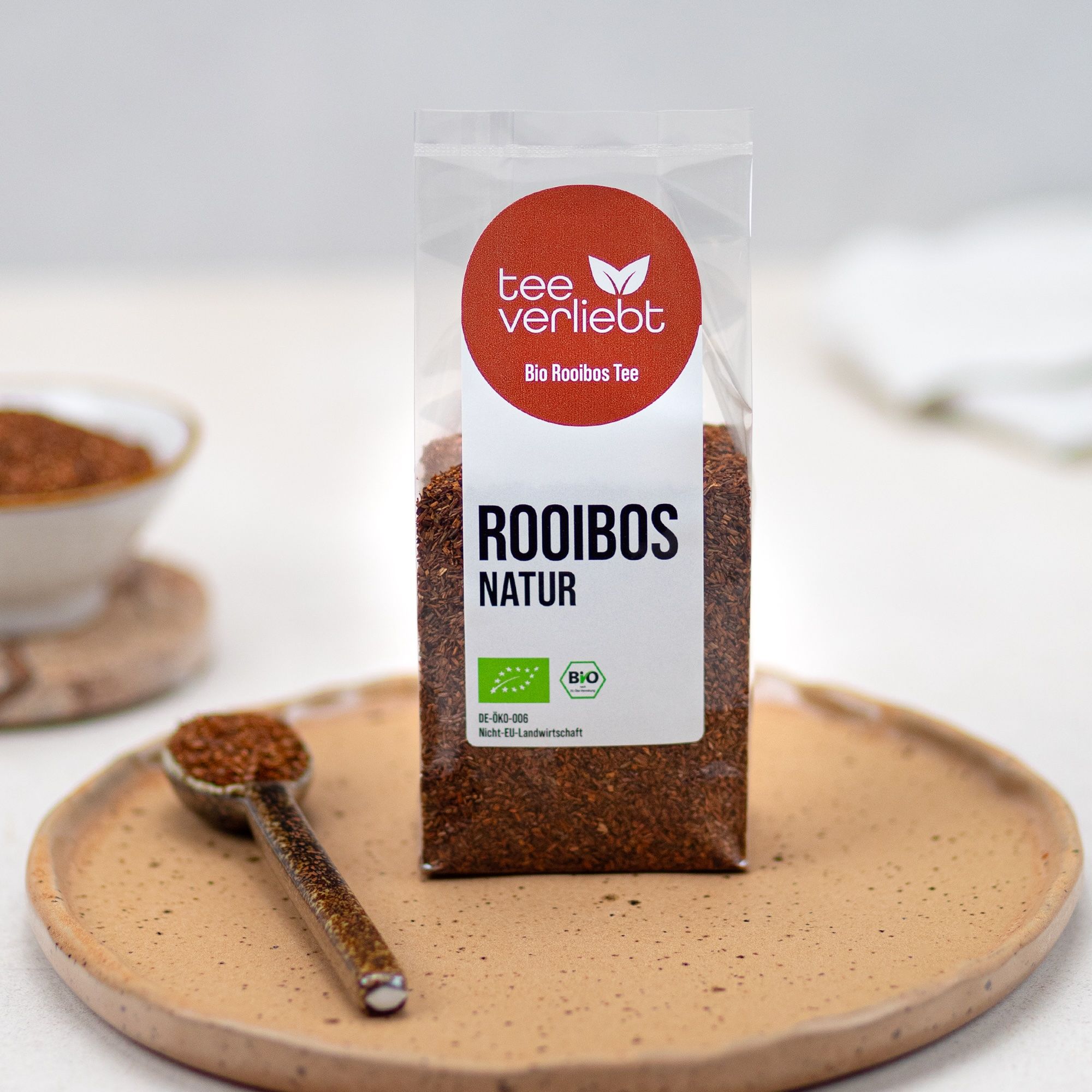 Verpackung mit losem Rooibos-Tee, daneben eine Schale und ein Löffel mit Tee. Aufkleber mit Logo und Text 'tee verliebt'.