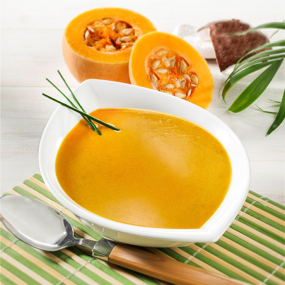 Schüssel Kürbissuppe, daneben Kürbisstücke. Auf Bambusmatte mit Löffel und Dekoration.