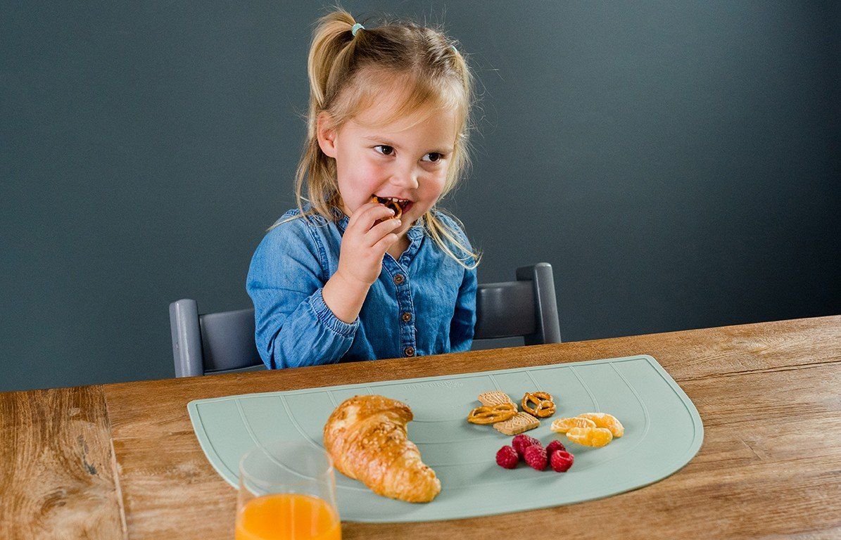 Kind isst am Tisch. Auf dem Platzdeckchen liegen Croissant, Obst und Brezeln.