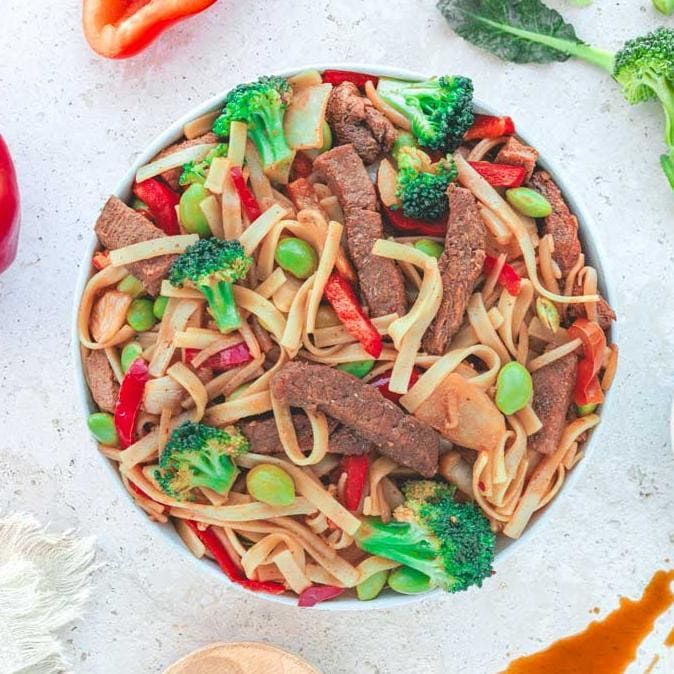 Nudelgericht mit Brokkoli, Fleischstreifen und Gemüse. Draufsicht. Auf hellem Untergrund. Rote Paprika und Soße.