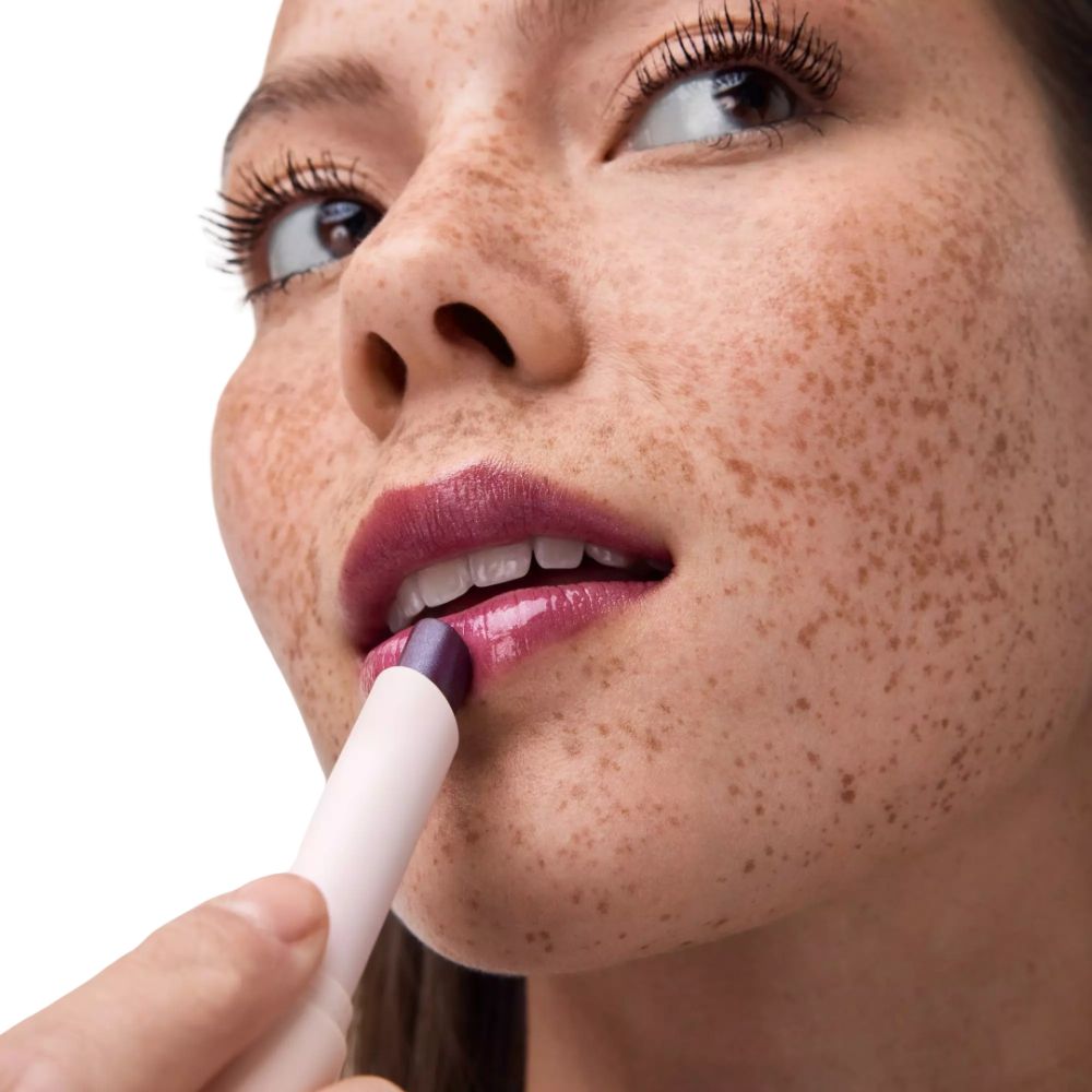 Frau mit Sommersprossen trägt lila Lippenstift auf.