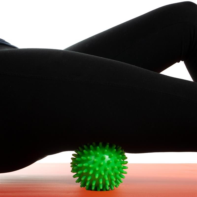 Grüner Massageball mit Noppen unter dem Bein einer Person, die auf einer Matte liegt.