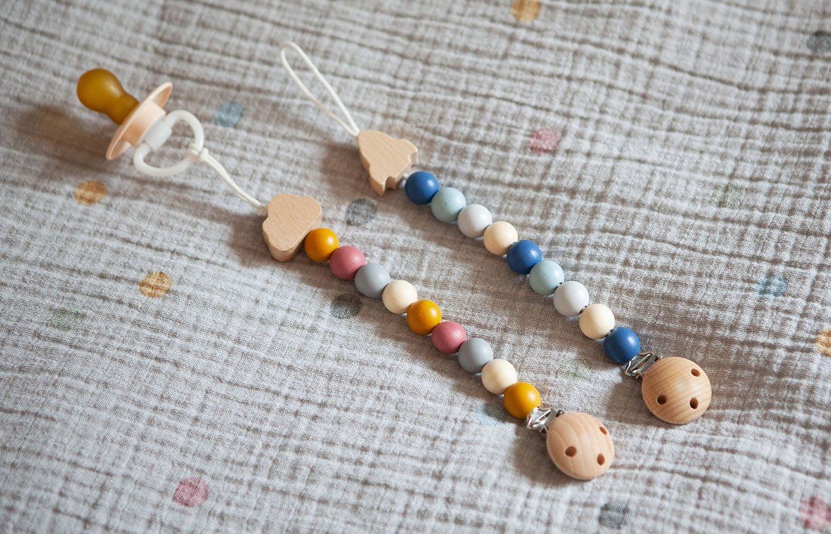 Zwei Schnullerketten mit Holzperlen, Clip und Holzfiguren. Eine mit Schnuller. Auf Stoffuntergrund.