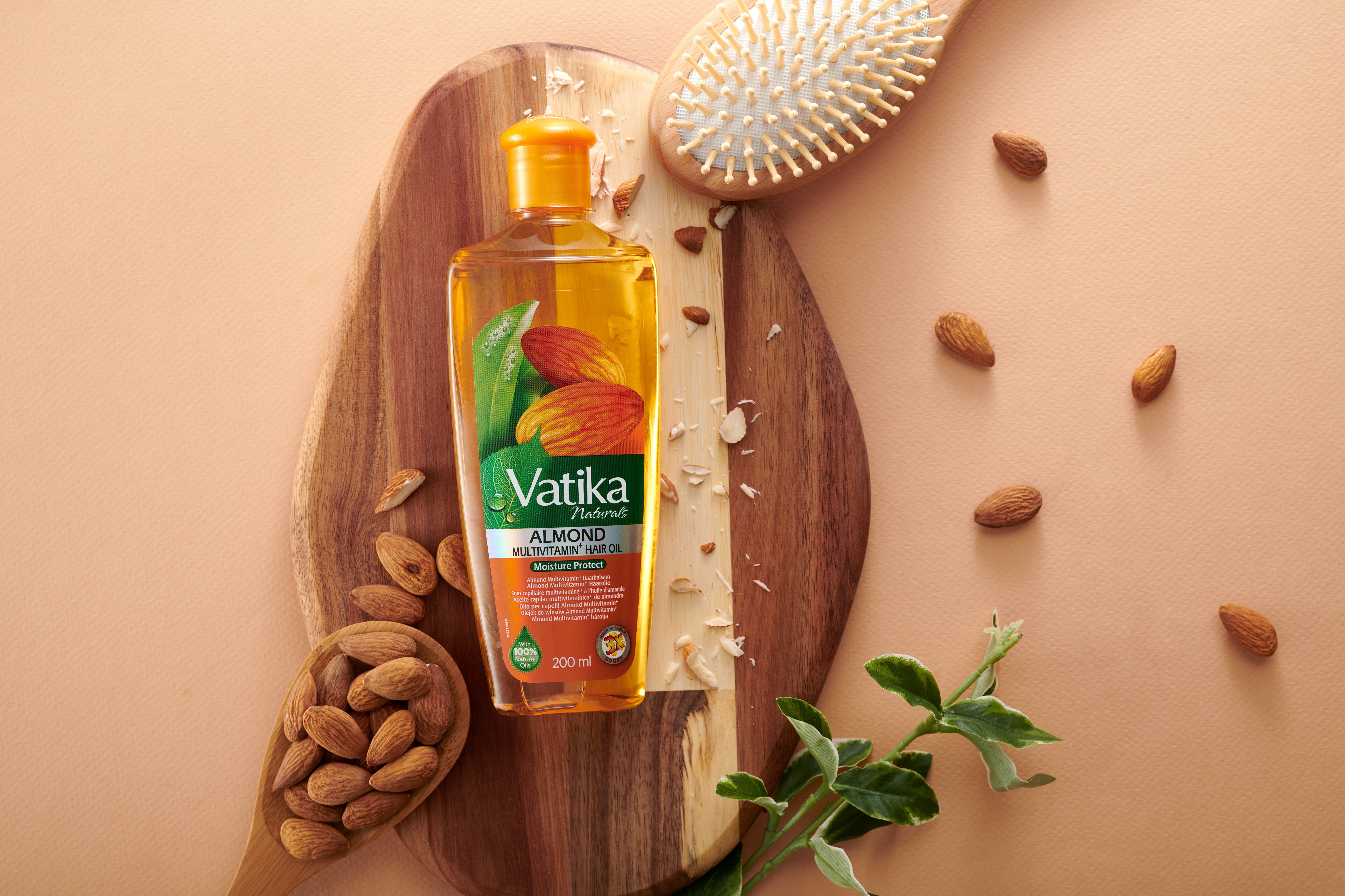 Flasche mit Haaröl auf Holzbrett, umgeben von Mandeln, Haarbürste und Blättern. Aufschrift: Vatika Almond Multivitamin Hair Oil.