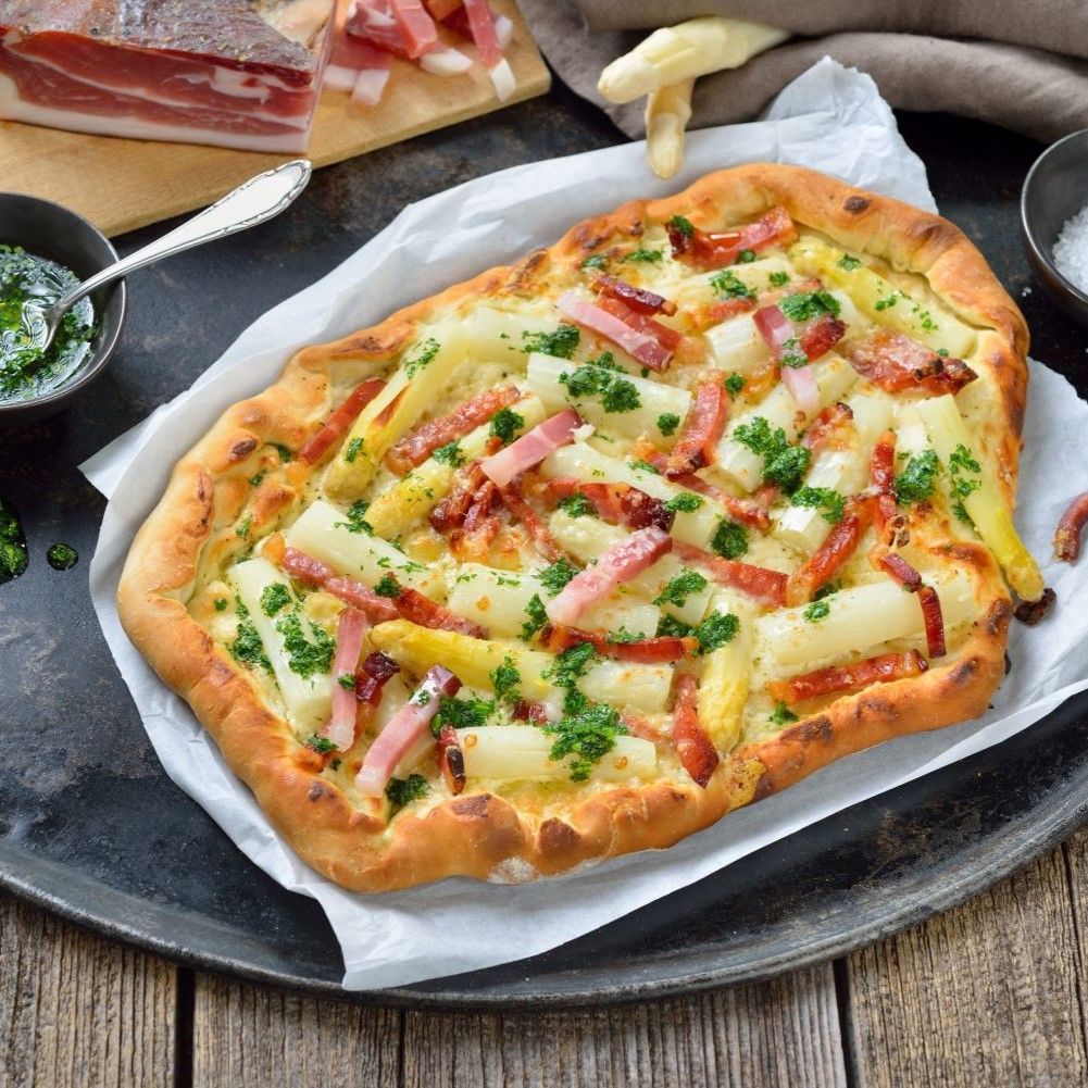 Großer Flammkuchen auf Tablett. Belegt mit Spargel, Speck und Petersilie. Dazu Schalen mit Soße und Salz.