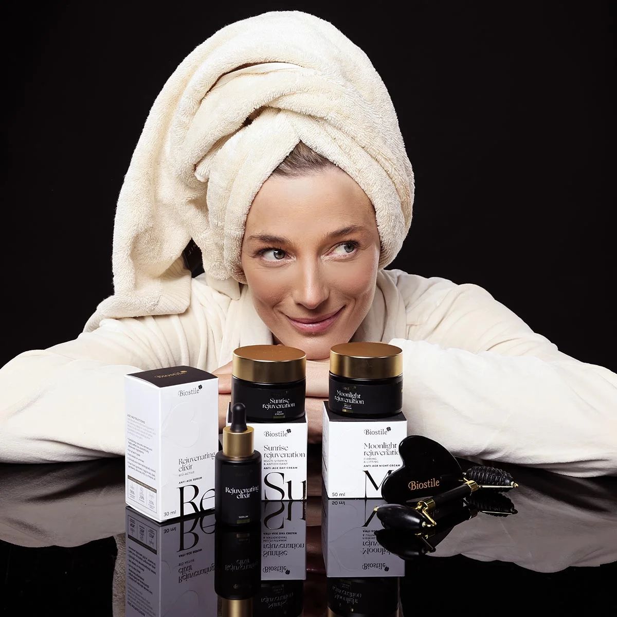 Donna con asciugamano e accappatoio, circondata da prodotti. Prodotti: crema, siero, strumento Gua Sha e rullo facciale.