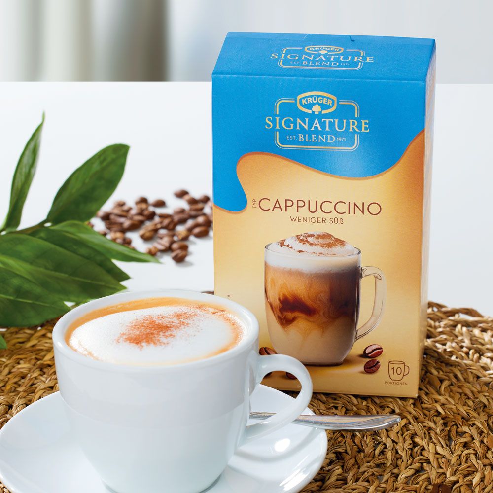 Krüger Signature Blend Cappuccino, weniger süß, in Verpackung. Cappuccino in Tasse daneben.