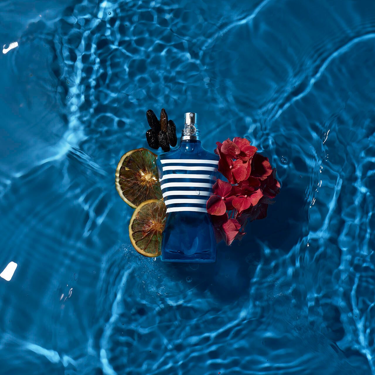 Blaues Parfümflakon in Torsoform in Wasser. Dazu: Blüten, Orangenscheiben, dunkle Blätter. Blaues Wasser.