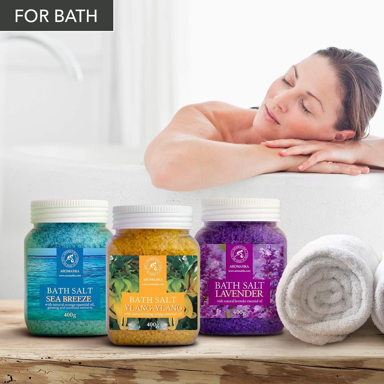 Drei Gläser Badesalz neben Badewanne. Gelb, blau und lila. Etiketten mit Produktnamen und Logo. Gelb: Ylang-Ylang. Blau: Meeresbrise. Lila: Lavendel. Handtücher.