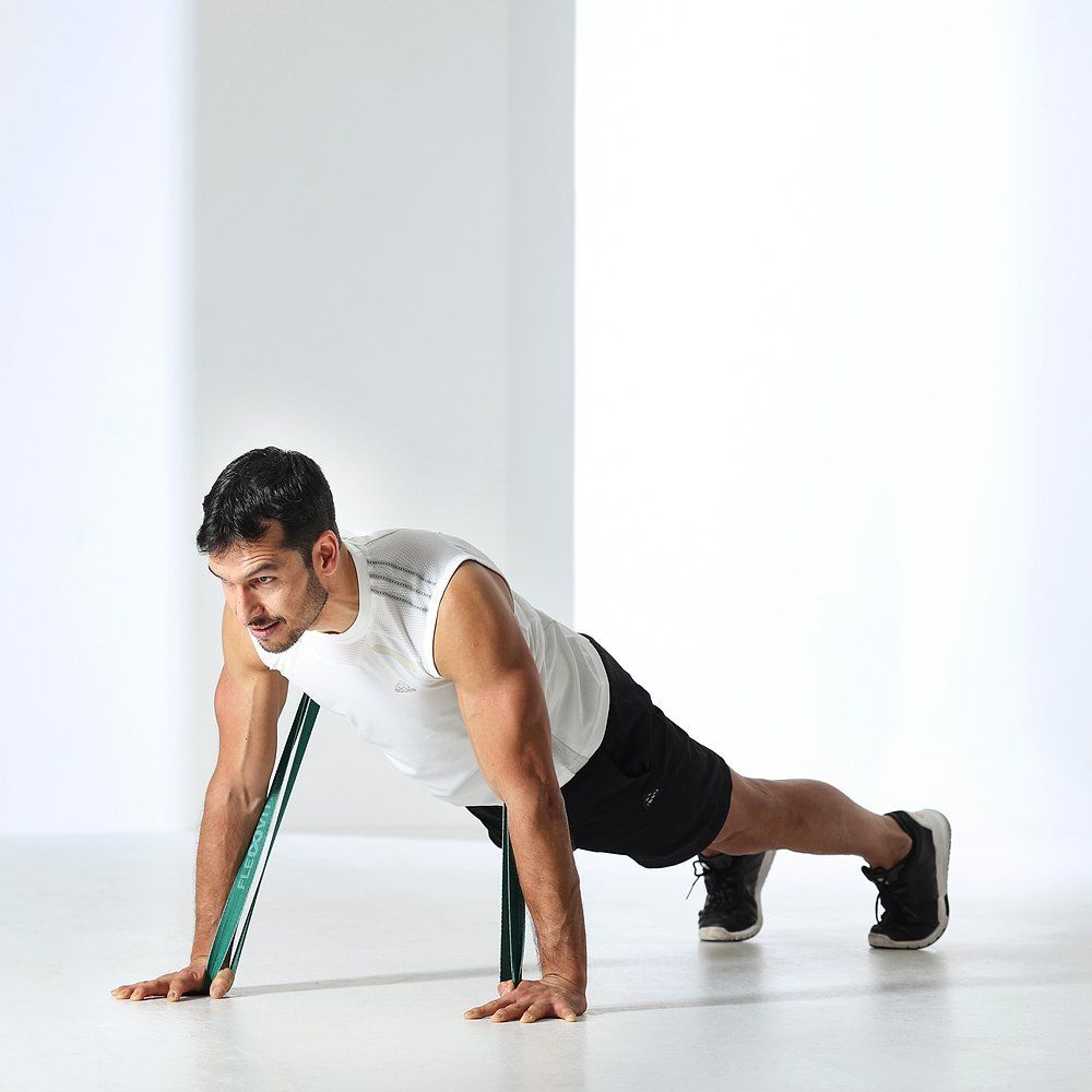 Mann macht Liegestütze mit grünem Fitnessband. Das Band ist um die Hände gelegt. Schwarze Shorts, weißes Tanktop.