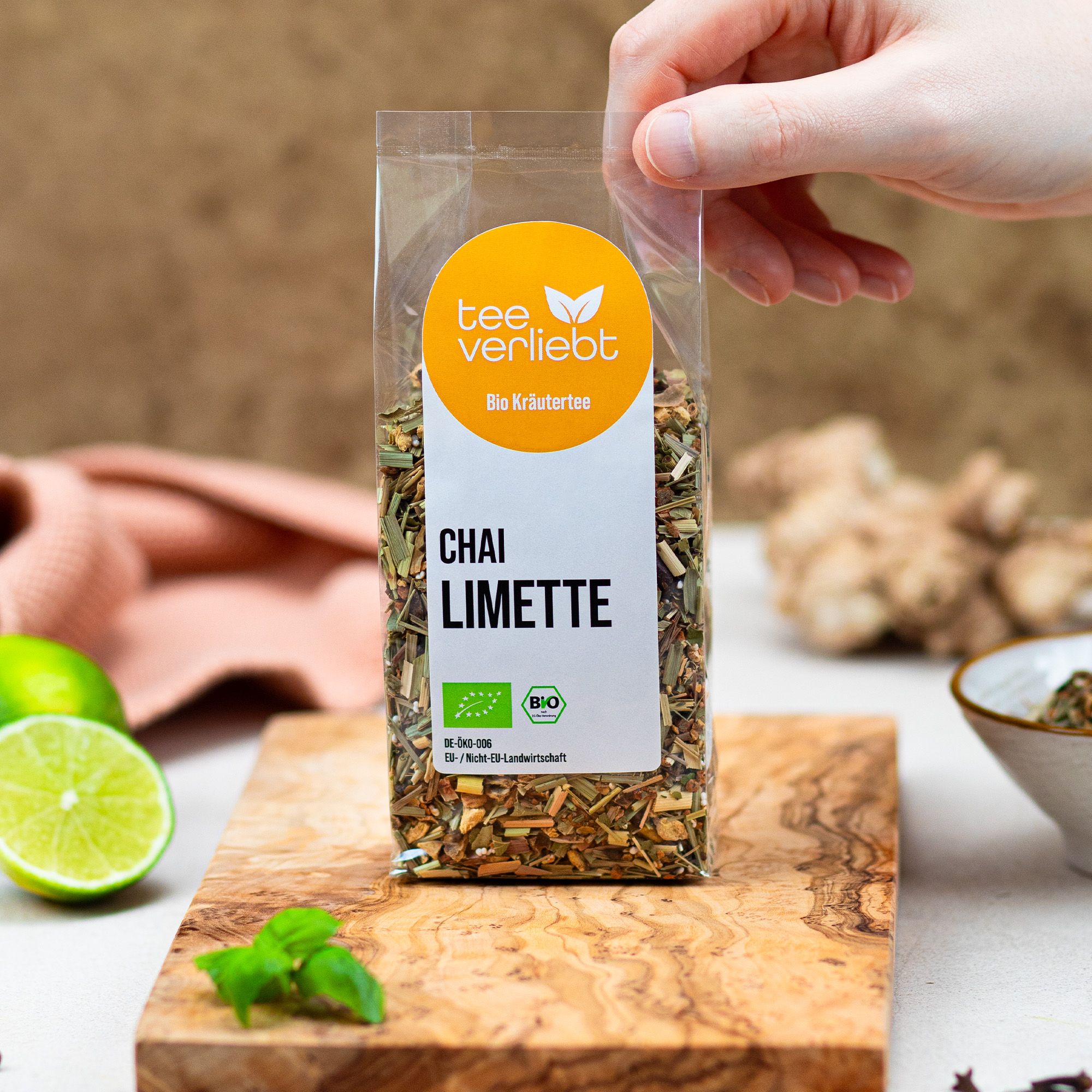 Hand hält transparente Tee-Verpackung. Aufkleber mit Logo, 'Chai Limette' und Bio-Siegel. Auf Holzbrett.