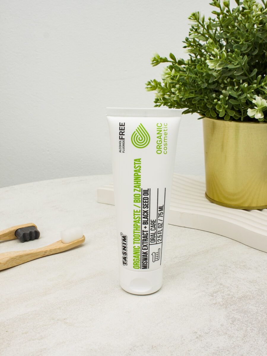 Weiße Zahnpasta-Tube mit grüner Grafik und Text. Zwei Bambus-Zahnbürsten und eine Topfpflanze im Hintergrund.