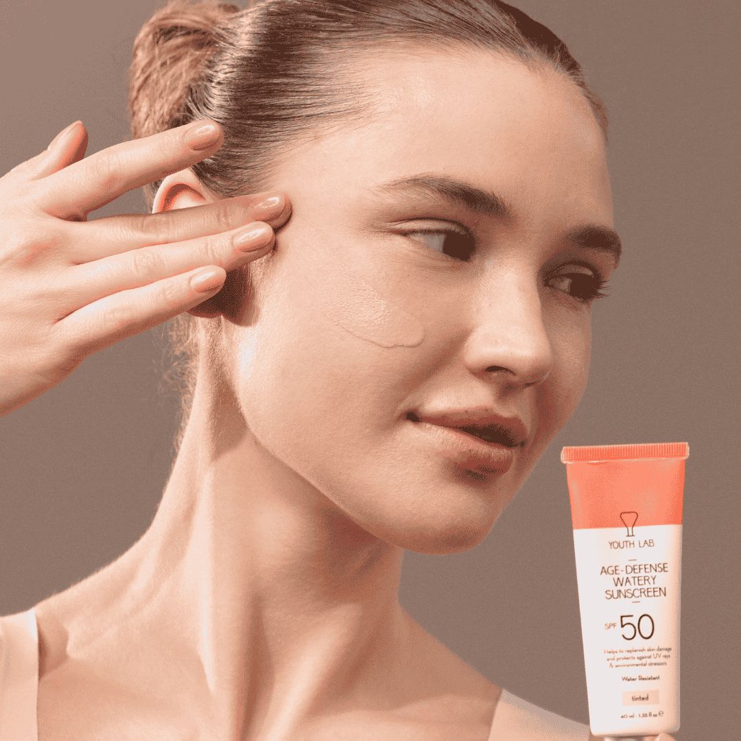 Frau trägt Produkt auf die Haut auf. YOUTH LAB Age-Defense Watery Sunscreen SPF50 Tinted Tube im Hintergrund. Produktname und SPF50.