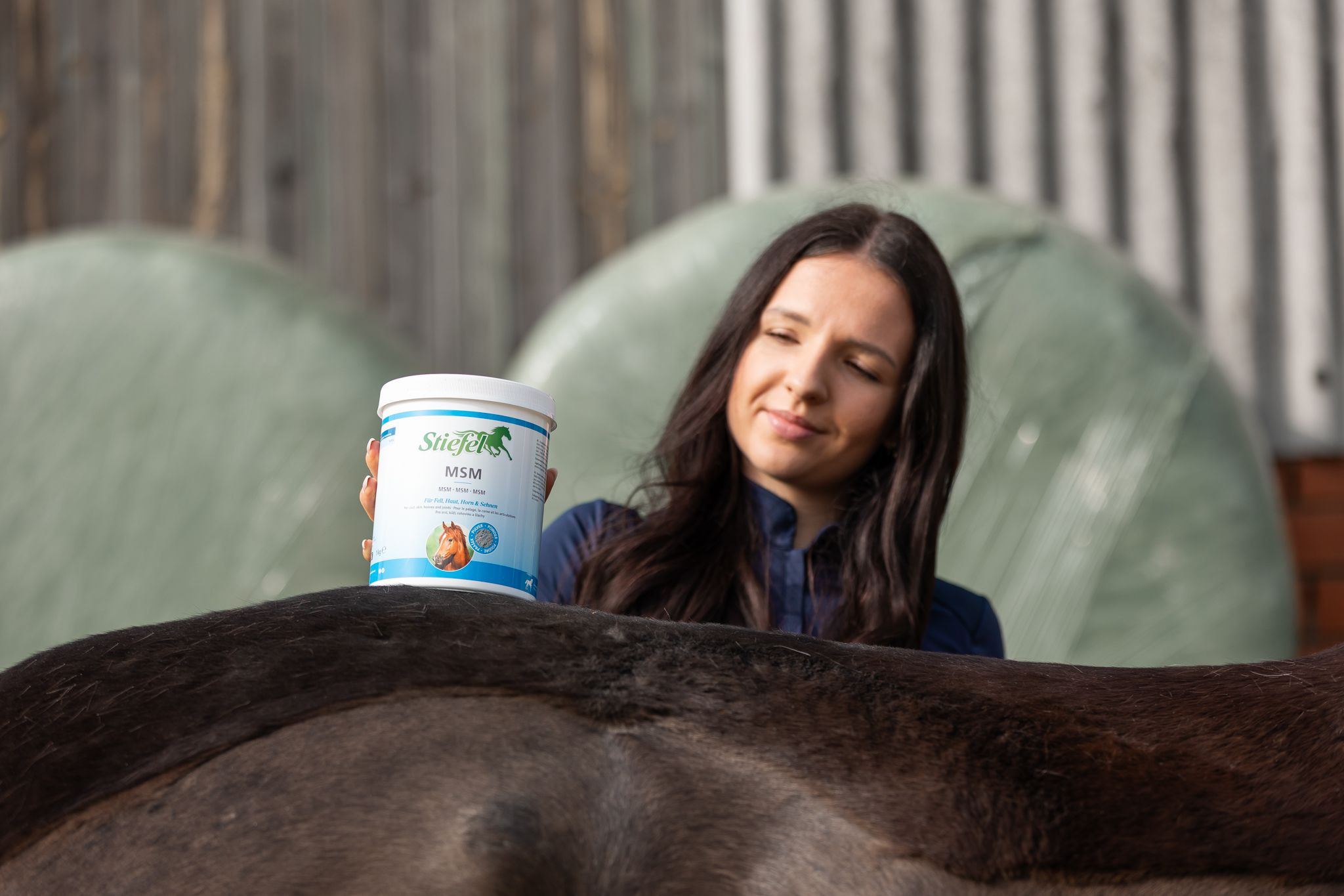 Frau hält einen Behälter über dem Rücken eines Pferdes. Der Behälter hat ein Etikett mit Text und einem Logo.