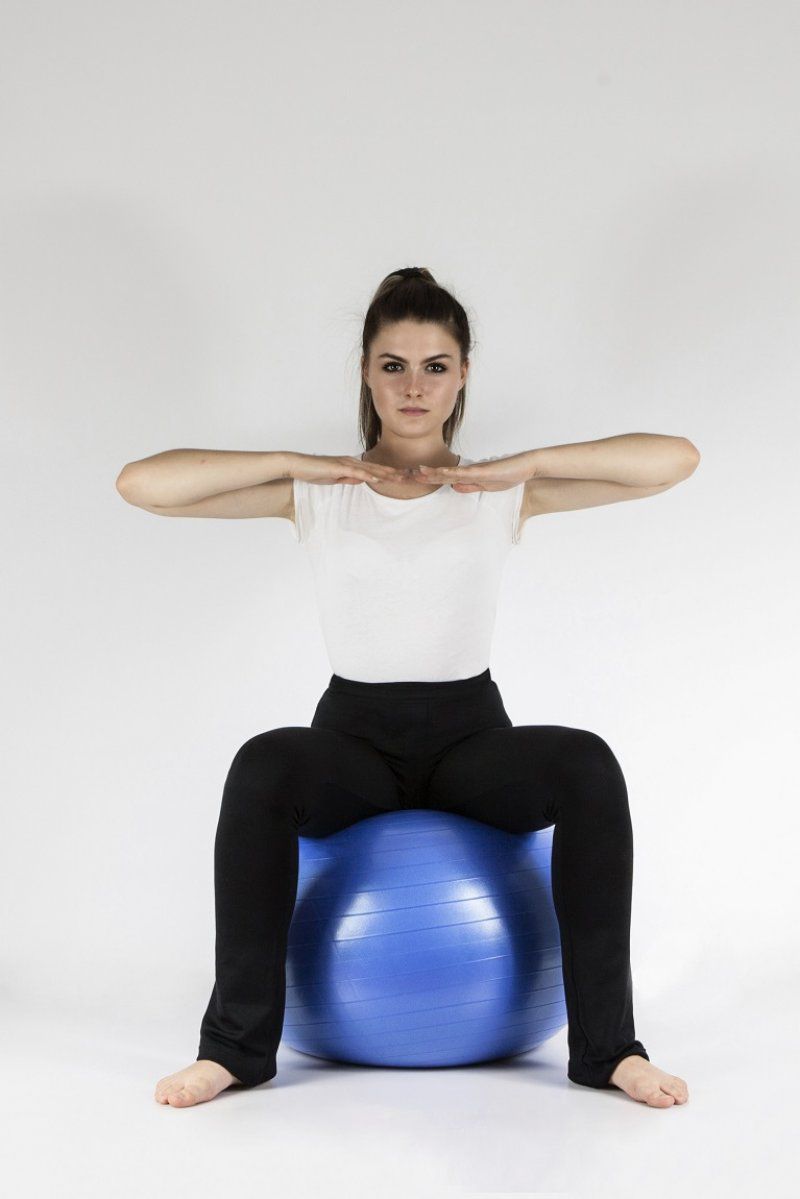 Frau sitzt auf blauem Gymnastikball, Arme horizontal ausgestreckt. Trägt schwarze Hose und weißes T-Shirt.