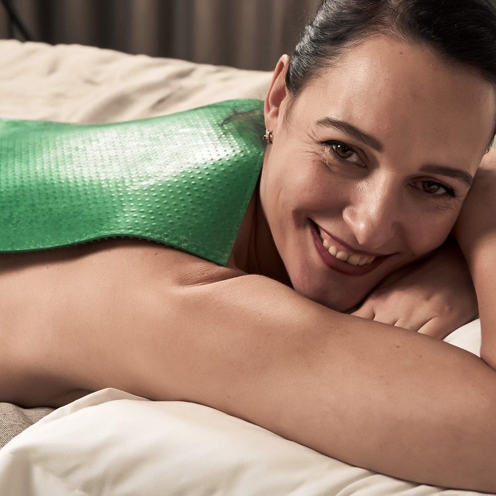 Frau liegt auf dem Bauch, ein grünes Produkt auf dem Rücken. Sie lächelt in die Kamera.