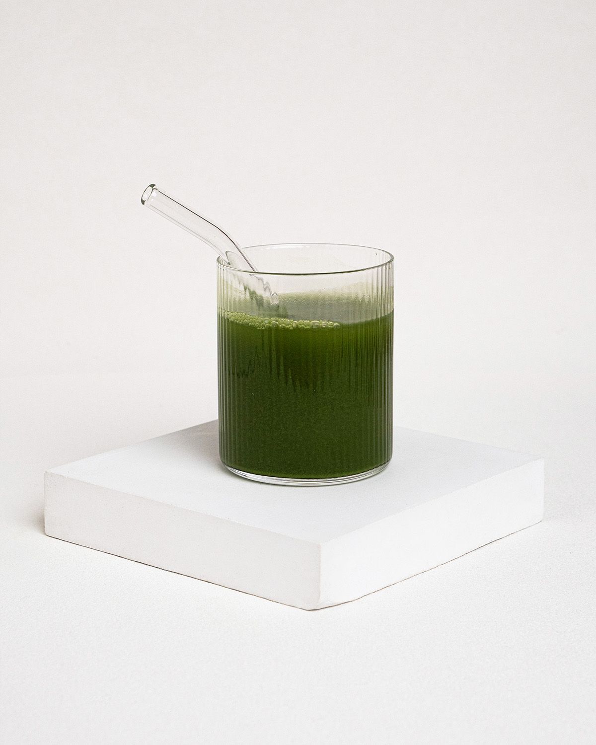 Glas mit grünem Getränk und Trinkhalm. Glas steht auf weißem Podest. Getränk ist grün.