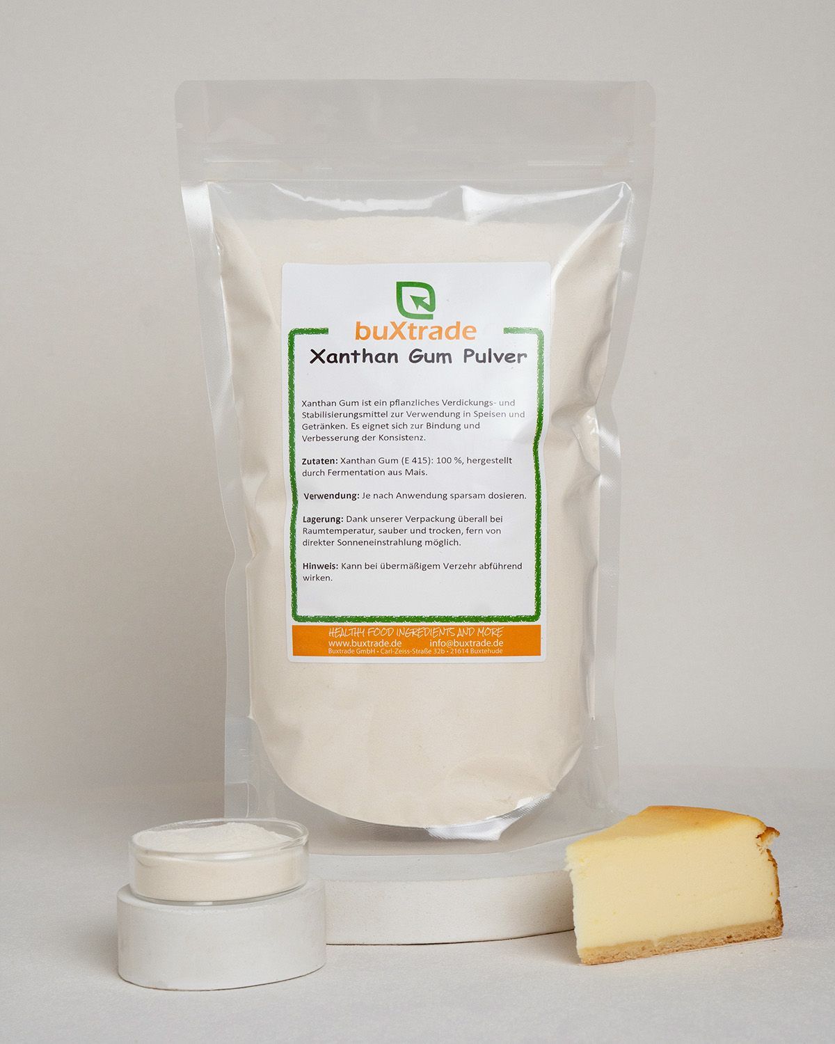 Verpackung mit buxtrade Xanthan Gum Pulver, daneben ein Glas mit Pulver, ein Löffel und ein Stück Kuchen.