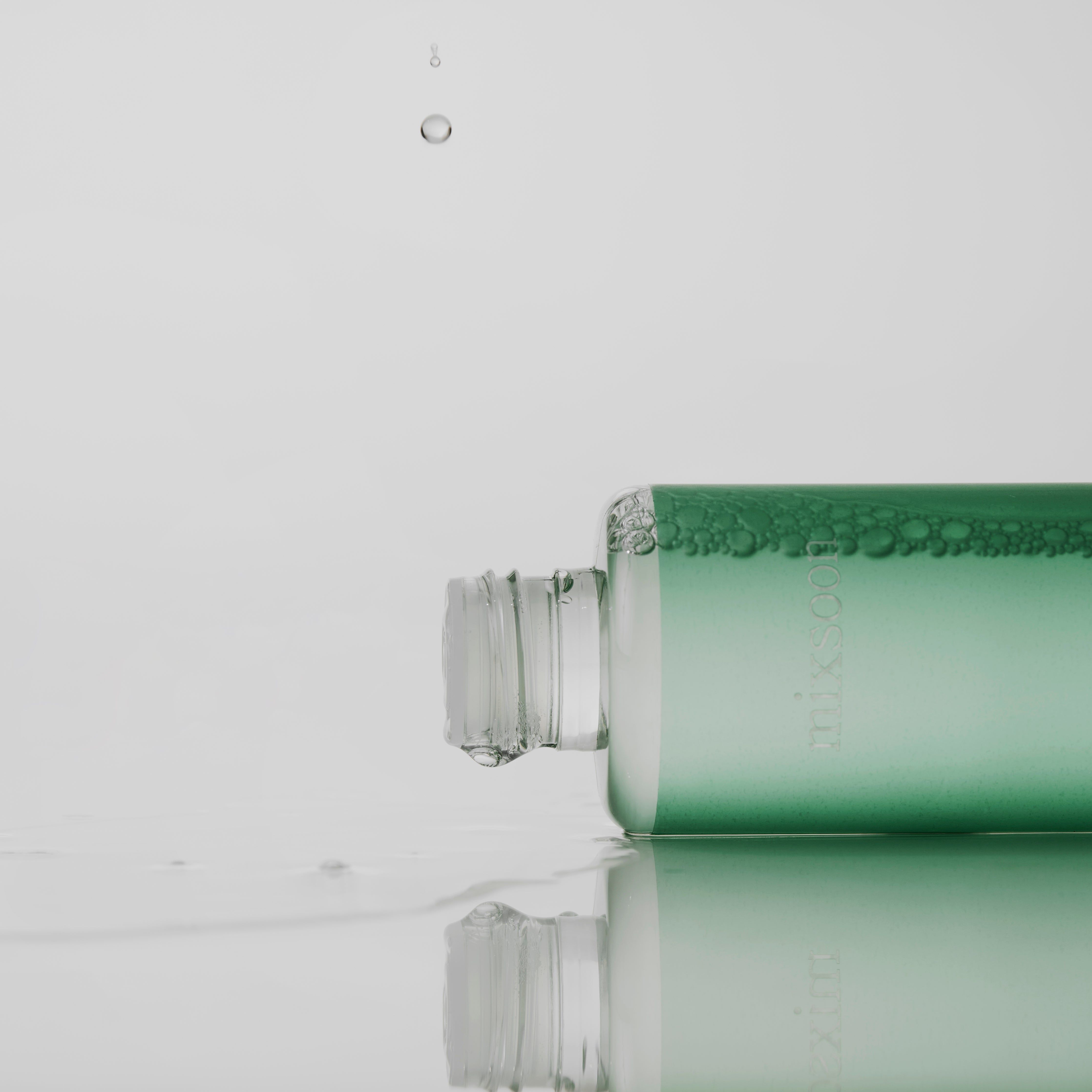 Grüne Flasche liegt. Aufschrift: mixsoon, Cicatree Clean Toner. Wassertropfen fallen. Hintergrund: weiß.