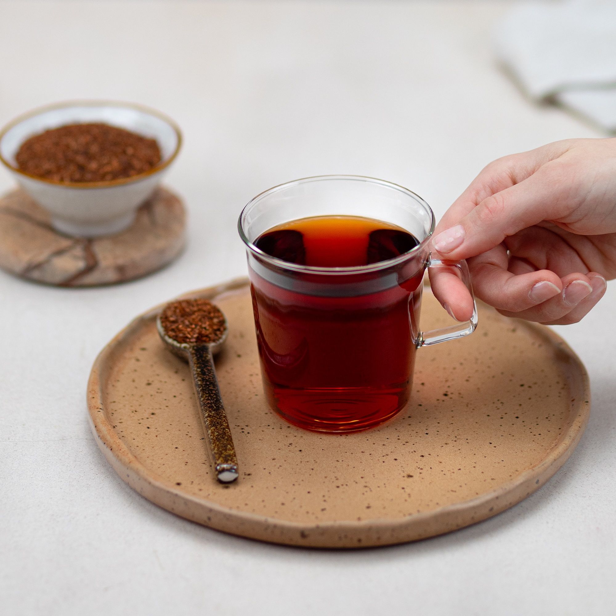 Glasbecher mit rotem Tee, daneben eine Schale und ein Löffel mit losem Tee. Hand hält den Becher. Auf einem Teller.
