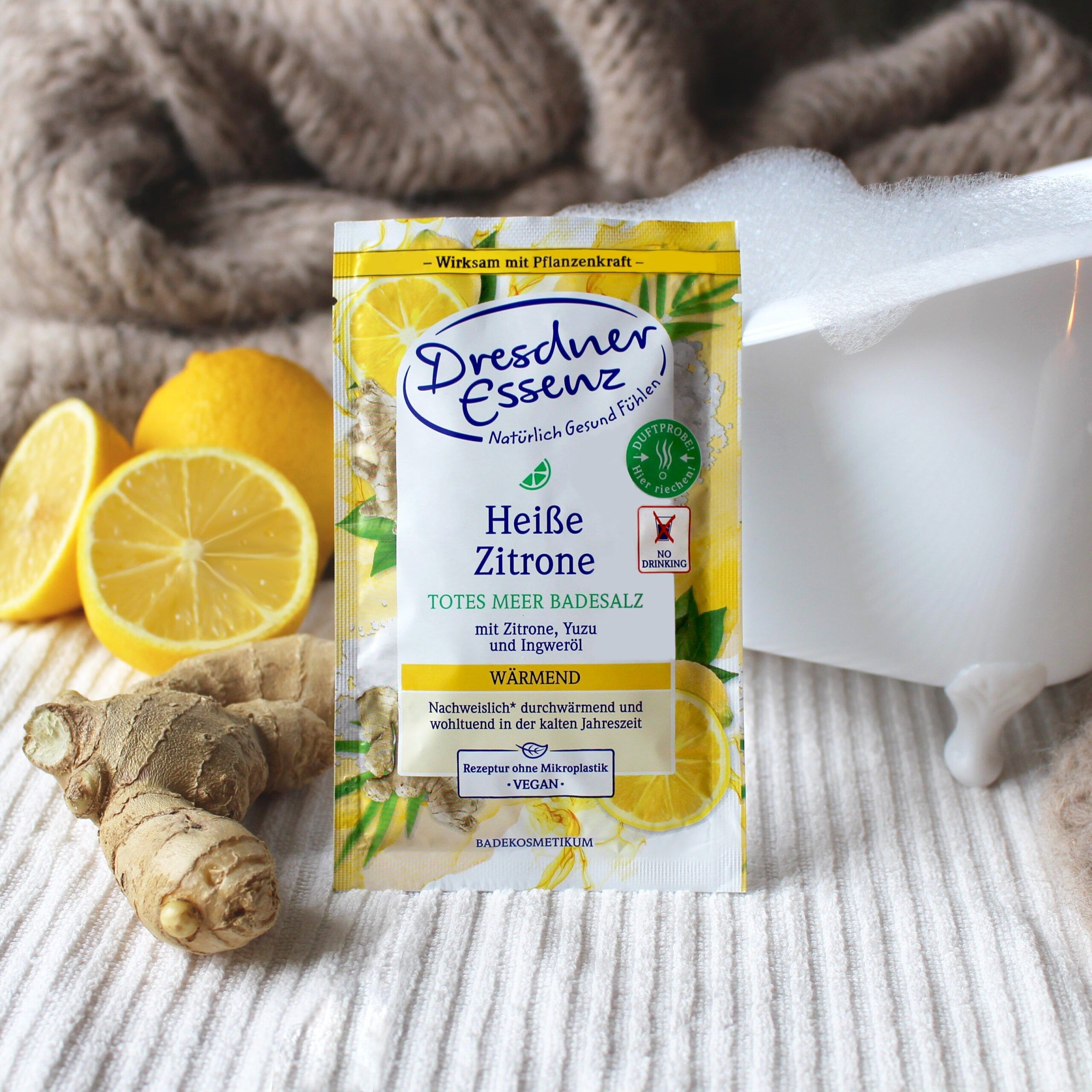 Verpackung von Dresdner Essenz Badesalz neben Zitronen, Ingwer und Badewanne. Aufschrift: Heiße Zitrone, Totes Meer Badesalz. Vegan.