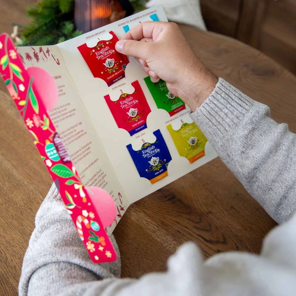 Person hält ein geöffnetes Tee-Buch. Zeigt Teebeutel. Roter Hintergrund mit floralem Dekor.