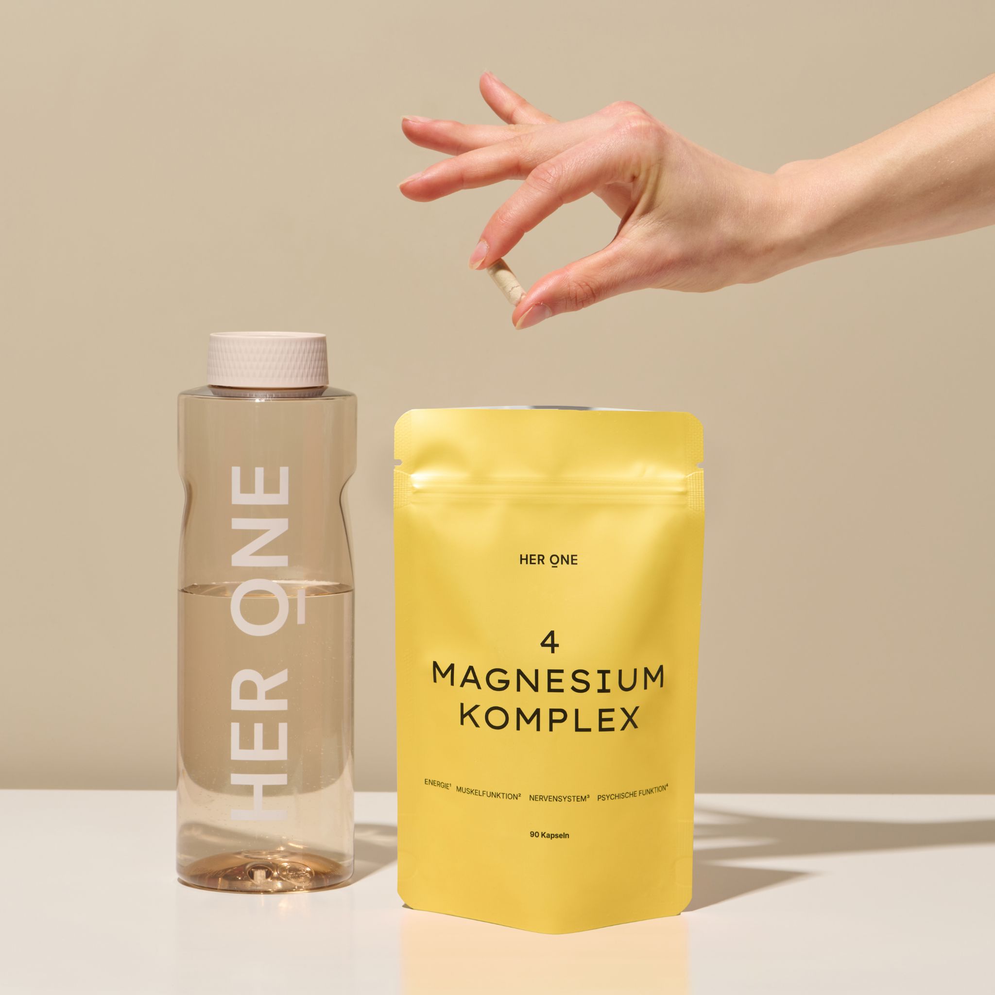 Gelbe Verpackung HER ONE 4 MAGNESIUM KOMPLEX neben Wasserflasche. Hand hält Kapsel.