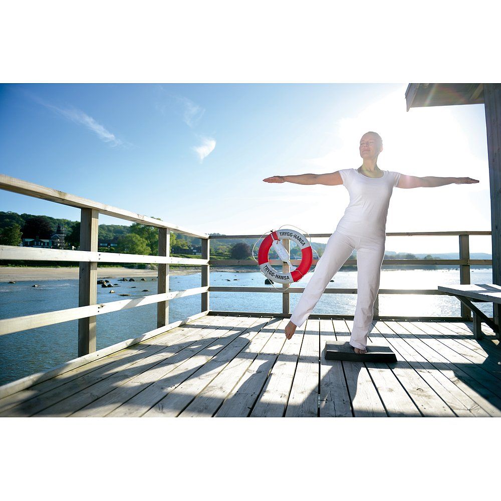 Person balanciert auf einem schwarzen Balance-Pad auf einem Steg. Meer und Himmel im Hintergrund.
