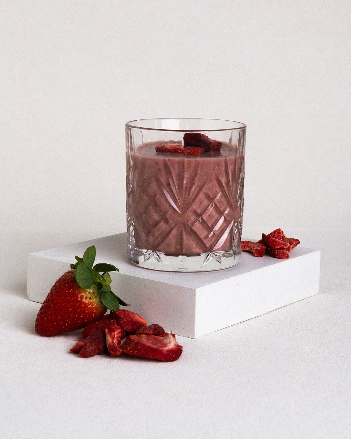 Glas mit Erdbeersmoothie auf weißem Podest. Frische Erdbeere und gefriergetrocknete Scheiben daneben.