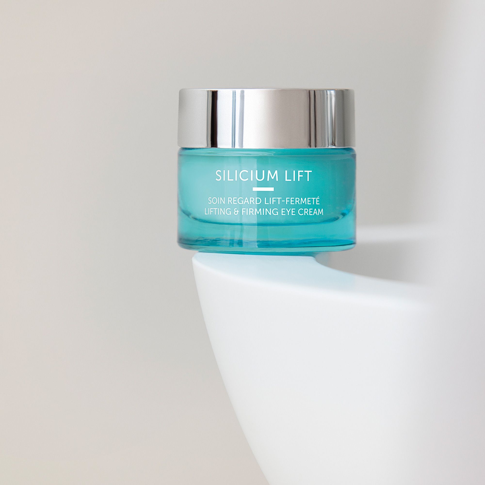 Türkisfarbener Tiegel mit silbernem Deckel. Aufschrift SILICIUM LIFT, darunter SOIN REGARD LIFT-FERMETÉ und LIFTING & FIRMING EYE CREAM. Auf weißem Podest.