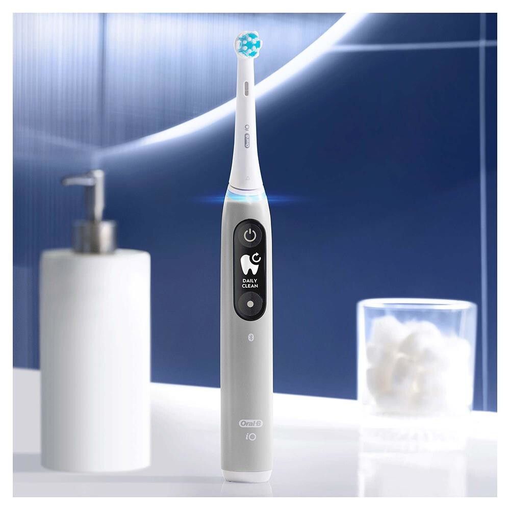 Graue elektrische Zahnbürste Oral-B iO Series 6 mit Ladestation. Im Hintergrund ein Waschbecken mit Seifenspender und Glas.