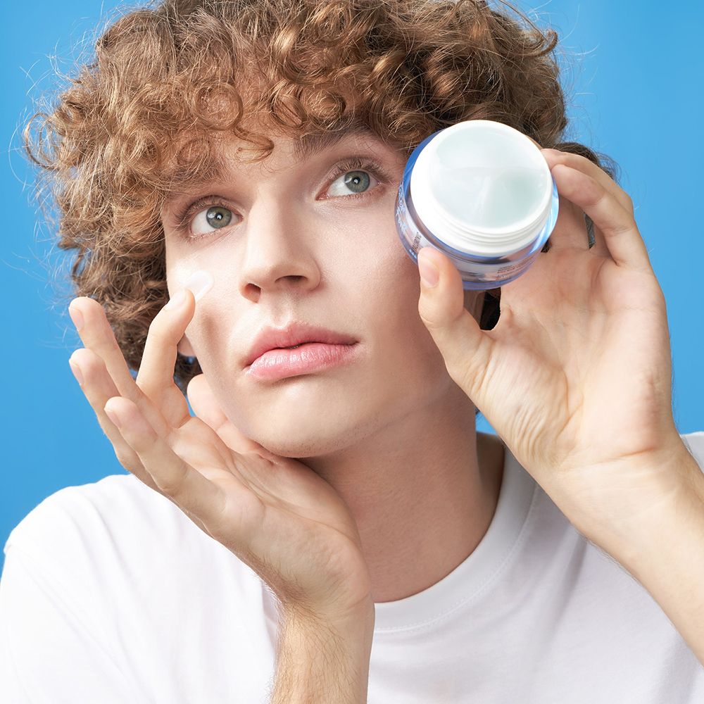 Mann hält Creme-Dose. Er berührt sein Gesicht. Blauer Hintergrund. Creme-Dose mit Aufschrift: Waterfull Hyaluronic.