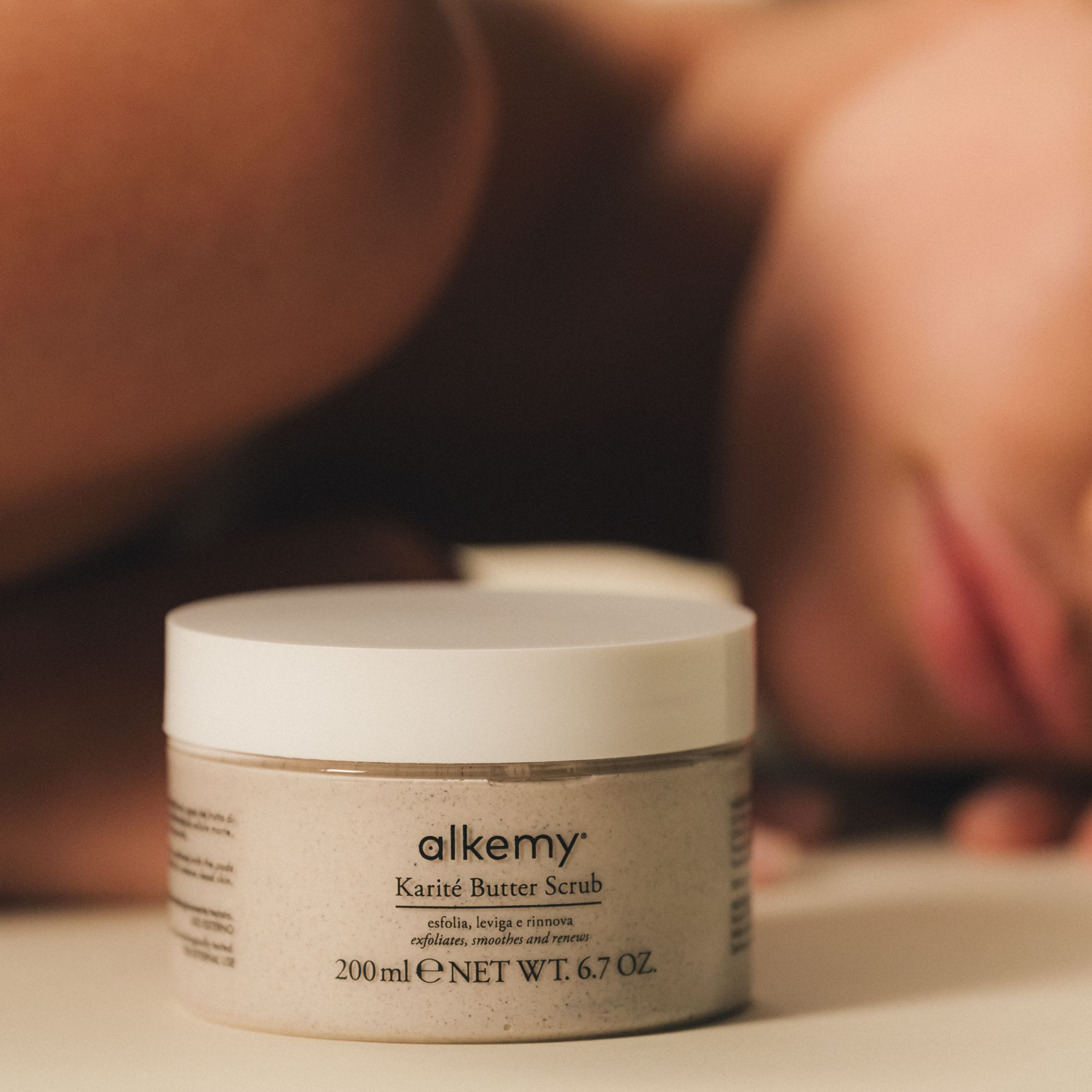 Alkemy Karité Butter Scrub Körperpeeling in einem Glasbehälter, neben einer liegenden Person.