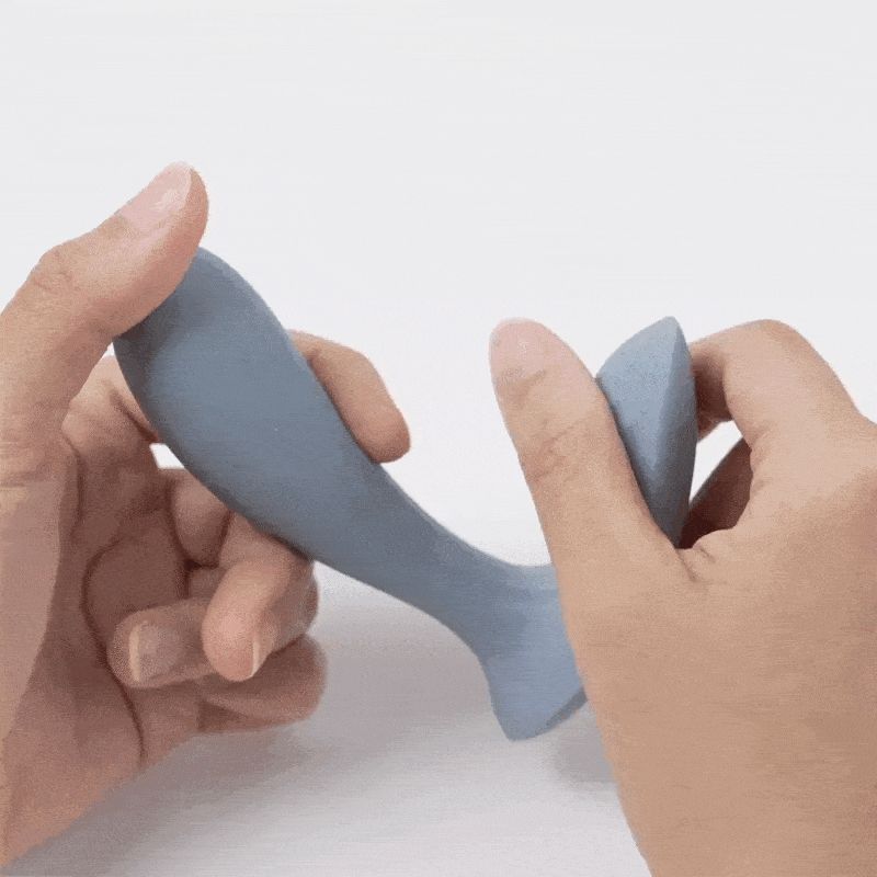 Graues Wellness-Produkt, in Händen gehalten. Ergonomische Form mit zwei abgerundeten Enden. Glatte Oberfläche.