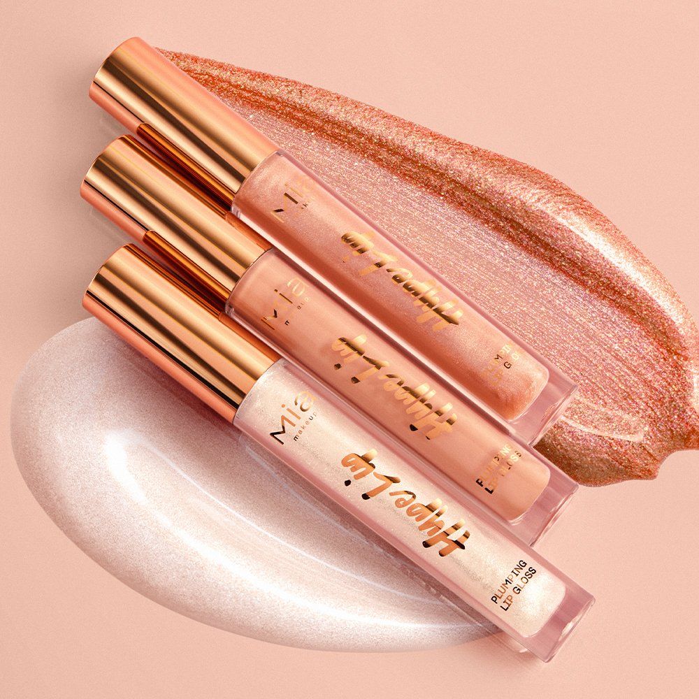Mia Make-up Hype Lip gloss volumizzante. Quattro lucidalabbra in diverse tonalità sono posizionati su una superficie rosa. I flaconi sono trasparenti con tappi dorati. C'è una macchia di colore rosa sulla superficie.