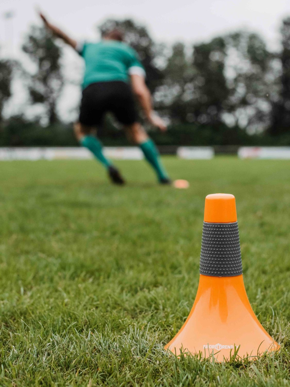 Orangener Speed Cone auf Rasen. Im Hintergrund ein Fußballspieler.